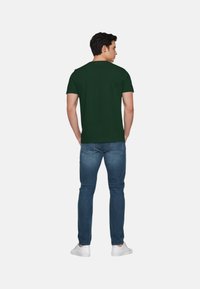 T-shirt verde scuro a maniche corte in cotone, abbinato a jeans in denim blu e sneaker bianche, mostrato dalla vista posteriore.