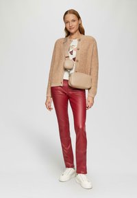 Beige gestrickte Cardigan, cremefarbener Oberteil mit geometrischem Druck, rote Lederhosen und weiße Sneaker. Außerdem enthält es eine Umhängetasche mit zwei Fächern.