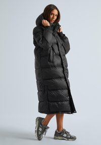 Melna garā pūkainā mēteli ar augstu apkakli un priekšējiem pogām, ar lielām kabatām un pāršūtu tekstūru. Apvienots ar pelēkiem sporta apaviem.