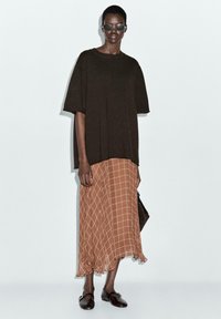 Dunkelbraunes, übergroßes T-Shirt, fließender brauner Rock mit Karomuster, legere braune Schuhe und runde Sonnenbrille, vor einem neutralen Hintergrund.
