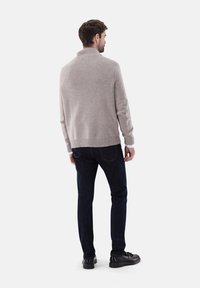 Beiger, strukturierter Strickpullover mit hohem Kragen, kombiniert mit dunklen blauen Jeans und schwarzen Schuhen, von hinten betrachtet.