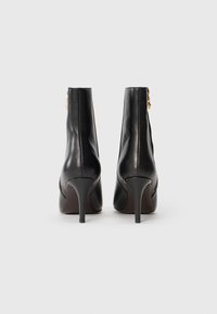 Bottines en cuir noir avec un bout pointu, un haut talon aiguille et un détail en métal doré sur le côté. Texture lisse et design épuré.