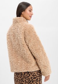 Bardzo puszysta kurtka z sztucznego futra w kolorze beżowym, z wysokim kołnierzem, luźnym krojem i długimi rękawami, połączona z brązową spódnicą w kratę w wzór lamparta.