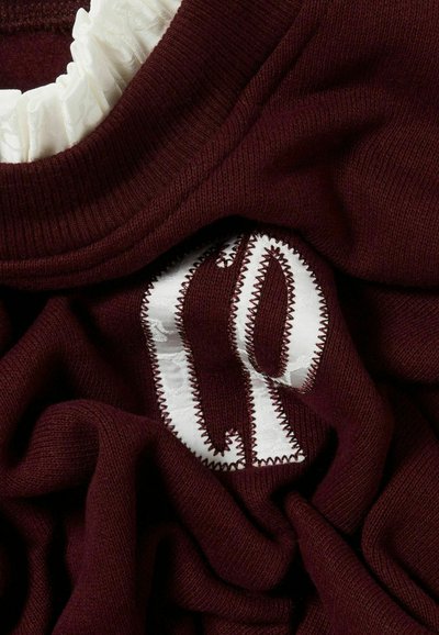 Suéter burdeos con un cuello blanco fruncido y iniciales bordadas en blanco, con un punto en zigzag alrededor de las letras. Textura suave.