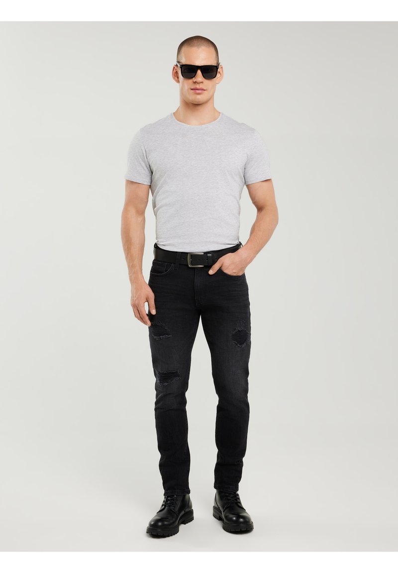 Mand iført sorte solbriller, lysegrå tætsiddende t-shirt, sorte revnede jeans, sort bælte og sorte støvler, stående mod en ensfarvet baggrund.