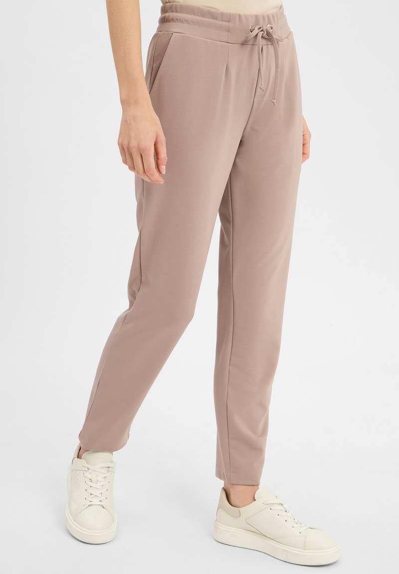 Jasnobrązowe dresy z elastycznym pasem i sznureczkiem. Gładki materiał, slim fit, kieszenie boczne. W zestawie z białymi sneakersami.