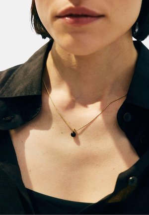 Primo piano del collo e delle labbra di una persona che indossa una maglietta nera e una sottile collana d'oro con un piccolo pendente nero.