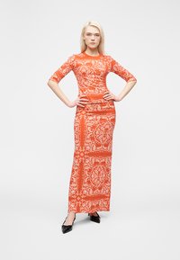 Γυναίκα που στέκεται με τα χέρια στη μέση, φορώντας ένα εφαρμοστό πορτοκαλί μάξι φόρεμα με λευκά μοτίβα παϊέτας και μαύρα μυτερά τακούνια.