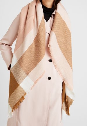 Femme portant une grande écharpe à carreaux beige et marron sur un trench-coat rose pâle avec des boutons noirs et un col roulé noir.