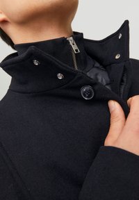 Abrigo de mezcla de lana negro con un cuello alto, cierre frontal con cremallera y botones a presión. Presenta un forro interior suave y una tela texturizada.