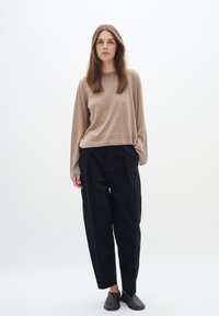 Maglione beige a maniche lunghe, vestibilità ampia. Pantaloni neri a gamba larga con pince. Calzature scure, design minimalista su uno sfondo bianco semplice.