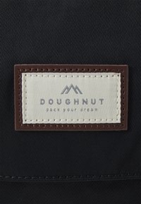 Czarna metka z logo "DOUGHNUT" oraz grafiką góry. Kremowe tło, brązowa obwódka i jasnobrązowe akcenty szwowe. Tekst brzmi "spakuj swoje marzenia".