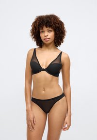 Juodas apatinis trikotažas, apimantis permatomą liemenėlę su minkštais puodeliais ir plačiomis petnešomis, derinamas su atitinkančiomis permatomomis aukšto liemens kelnaitėmis.