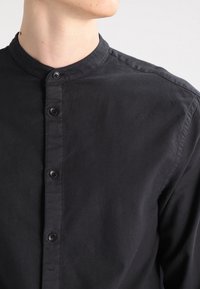 Camisa negra de botones con cuello de banda, que presenta una textura suave, un brillo sutil y seis botones negros a lo largo de la tapeta delantera.
