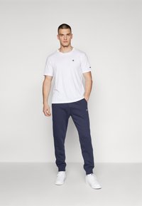 Weißes T-Shirt mit Rundhalsausschnitt und kleinem Logo, kombiniert mit marineblauen Sweatpants. Beide aus weichem Stoff gefertigt. Modell trägt weiße Sneakers.
