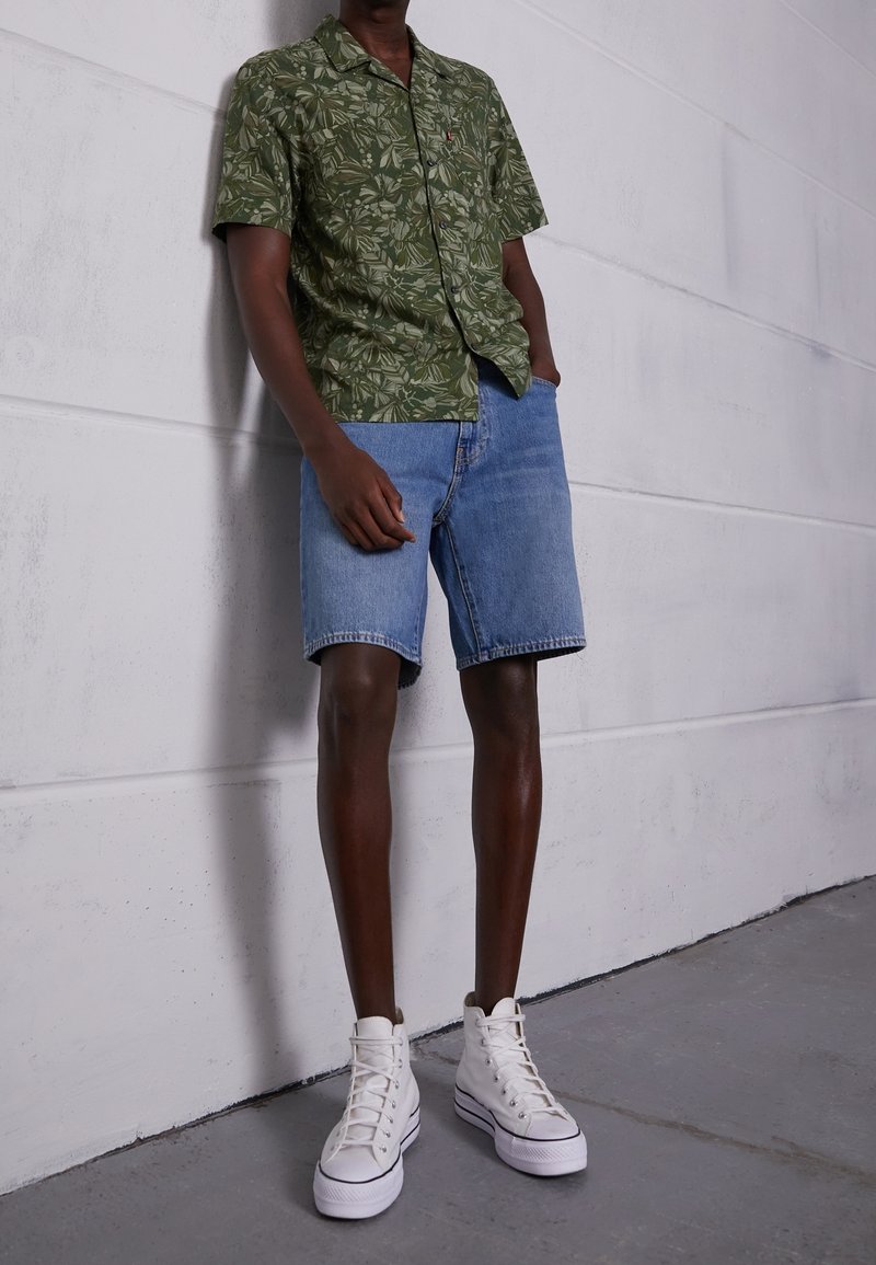 Chemise à manches courtes vert à motifs, accompagnée d'un short en denim bleu avec ourlets retroussés et des baskets blanches montantes. Fond mural de couleur neutre.