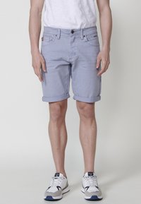 Vīrietis, ģērbies gaiši zilos sarullētos džinsu šortos, baltā T-kreklā un baltās kedās ar jūras zilām un pelēkām akcentiem, stāv pret vienkāršu fona.