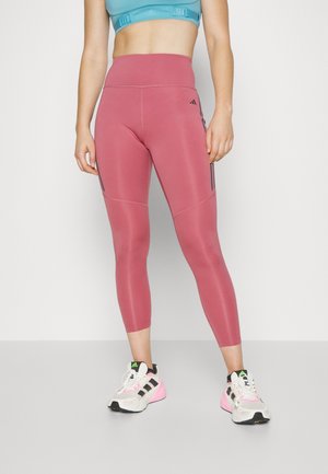 Leggings de sport taille haute, roses, avec des rayures noires sur les côtés, en matière extensible. Portés avec un crop top bleu clair et des baskets.