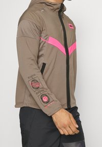 Bruine zip-up jas met een roze chevron ontwerp, textiel stof, een capuchon en grafische details op de mouwen, inclusief een smiley gezicht.