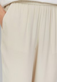 Pantalons beige à taille élastique avec poches latérales, présentant une texture lisse et une coupe ample. Le tissu semble léger et fluide.