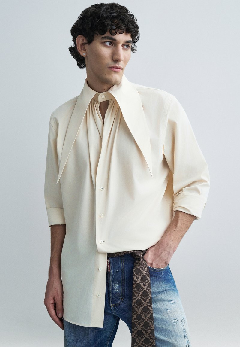 Cream-colored button-up shirt with oversized collar, featuring pleats, paired with distressed blue jeans and a patterned brown belt.