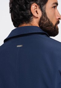 Homme aux cheveux bouclés foncés et barbe portant une veste bleu marine à col, vu de dos et de profil latéral.