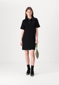 Melnā polo kleita ar īsām piedurknēm, rakstainu apkakli un izšūtu logotipu. Saskaņota ar melnām zeķēm un papēžu apaviem, turot zaļu somu.