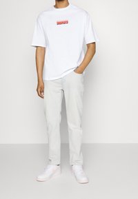 Bílá bavlněná tričko s červeným grafickým logem, světlé modré džíny a bílé tenisky s růžovými akcenty. Neformální outfit v postoji.