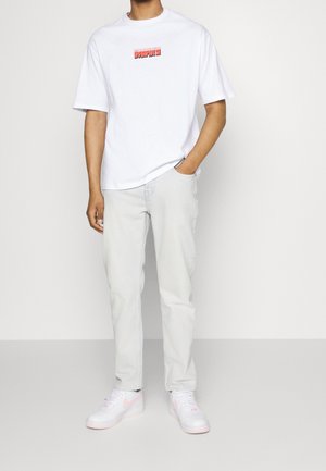 Camisa de algodón blanca con un logo gráfico rojo, jeans de mezclilla azul claro y zapatillas blancas con detalles en rosa. Pose de pie con un atuendo casual.