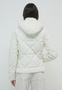 Frau, die eine weiße gesteppte Kapuzenjacke und helle Hosen trägt, steht mit einer Hand an der Hüfte, mit dem Rücken zum Betrachter.