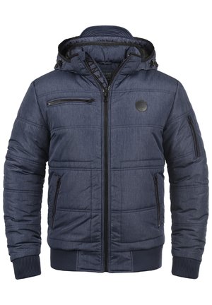 Homme portant une veste matelassée bleu marine avec plusieurs poches zippées et un jean gris, debout, une main dans la poche.