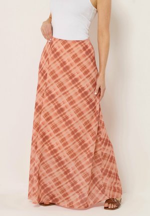 Femme portant une jupe longue fluide à carreaux rose et marron, associée à un haut blanc sans manches et des sandales marron.