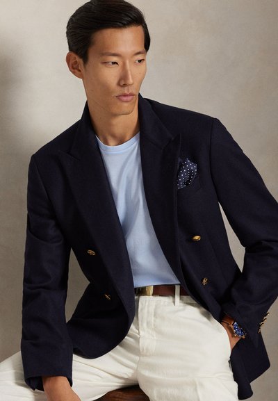 Άνδρας με διπλόμπαλο μπλέ σακάκι με χρυσά κουμπιά, ανοιχτό μπλε πουκάμισο, λευκό παντελόνι και μπλε μαντιλάκι τσέπης με πουά, καθισμένος με το ένα χέρι στην τσέπη.