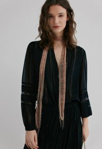 Blouse en chiffon noir à manches longues, avec des accents en dentelle et un décolleté en V profond, associée à une écharpe rose clair ornée de détails en perles.