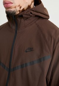 Brown Nike jacket with a black logo, featuring a hood, zip closure, and a horizontal black stripe across the chest, made from lightweight fabric.