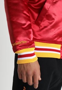 Červená saténová bunda s žluto-bílými pruhovanými žebrovanými lemy na rukávech. Na rukávu je žlutě vyšitá logo "Mitchell & Ness".