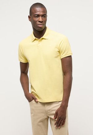 Man wearing yellow short-sleeve polo shirt and beige pants, standing with one hand in pocket against plain light background.