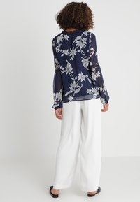 Blusa trasparente blu navy con ricami floreali bianchi, maniche lunghe con polsini elastici, abbinata a pantaloni bianchi a gamba larga e ballerine nere.