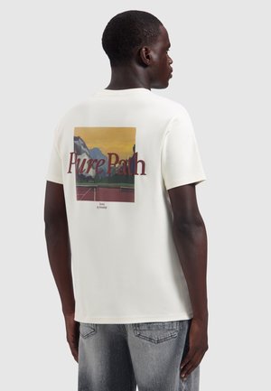 Man draagt een wit T-shirt met een afbeelding van een tennisbaan en bergen, tekst "Pure Path" op de achterkant, gecombineerd met vervaagde grijze jeans.