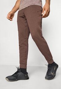 Mand iført brune joggebukser og sorte Nike sneakers står mod en hvid baggrund, benene let spredte.