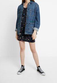 Farkkutakki nappikiinnityksellä, useilla taskuilla ja käärittyillä hihoilla mustan mekon päällä, jossa on värikäs lehtikuosi; yhdistetty mustiin tennareihin.