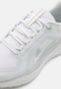 Vit idrottssko med ovandel i mesh och syntetmaterial, texturerat mönster, platta vita skosnören och grå accenter, med en synlig Nike-logga och Air Zoom-varumärke.