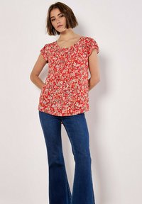 Top floral rojo con mangas cortas, detalles fruncidos y tela ligera, combinado con jeans oscuros acampanados.