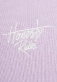 Lila stickad tyg med vit broderad text som säger "Ärligt talat regler," med en jämn textur och lätt ribbing.