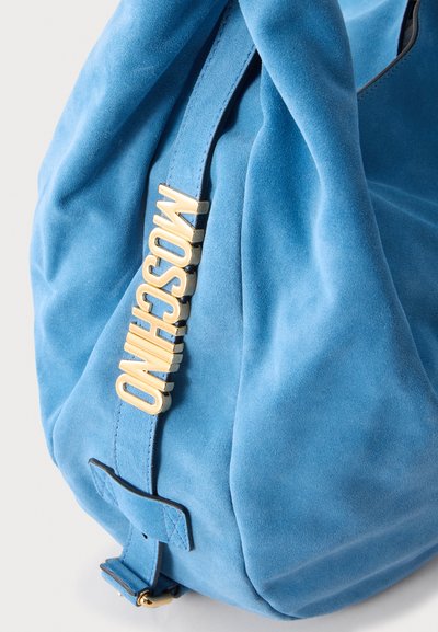 Sac en daim bleu avec un logo en métal doré portant l'inscription MOSCHINO fixé sur une bandoulière sur fond blanc.