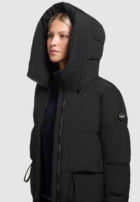 Giubbotto imbottito nero con un grande cappuccio, chiusura con zip, coulisse regolabili e un'etichetta rotonda con logo sul maniche sinistro.