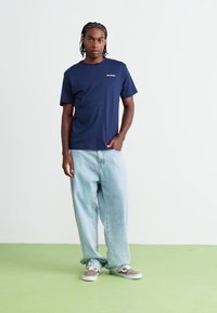 Marineblå kortærmet t-shirt med lille logo på brystet, parret med lyseblå løstsiddende jeans og grå sneakers på et grønt gulv.