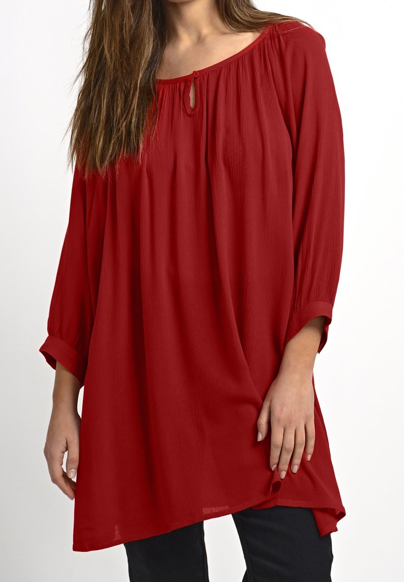 Femme portant une tunique rouge à manches longues ample avec un col fendu et un pantalon noir sur un fond blanc.