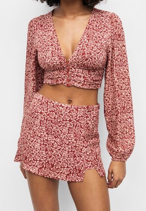 Femme portant un crop top rouge à fleurs à manches longues avec un décolleté plongeant en V et une mini-jupe taille haute assortie avec une petite fente à l'avant.