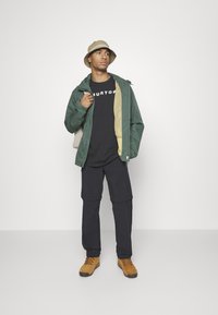 Groene waterdichte jas met beige voering, zwart t-shirt, zwarte cargo pants en bruine laarzen. Het model draagt een beige bucket hat.